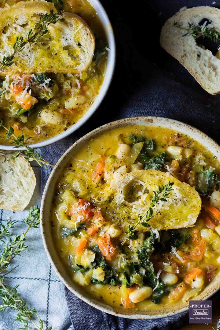 overhead view of ribollita soup served in 2 bowls and topped with thyme and parmesan Ribollita vegetable and bean soup served up in two bowls and topped with crusty bread, grated parmesan and fresh sprigs of thyme.